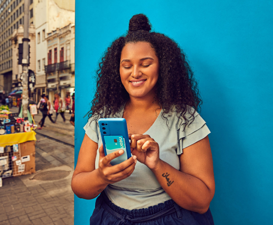 Mulher na rua sorrindo e mexendo em celular com cartão de crédito Neon na parte de trás do aparelho