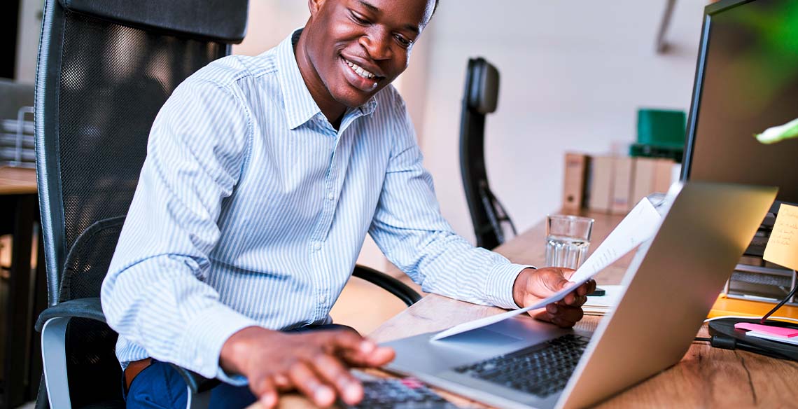 Homem sorrindo em frente a computador segurando papel em uma mão e usando calculadora na outra