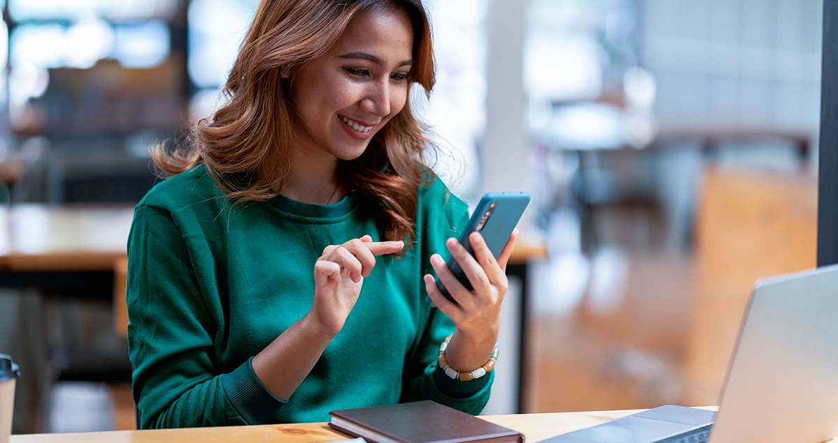 Mulher segurando o celular e sorrindo sentada em mesa com computador e caderno à sua frente