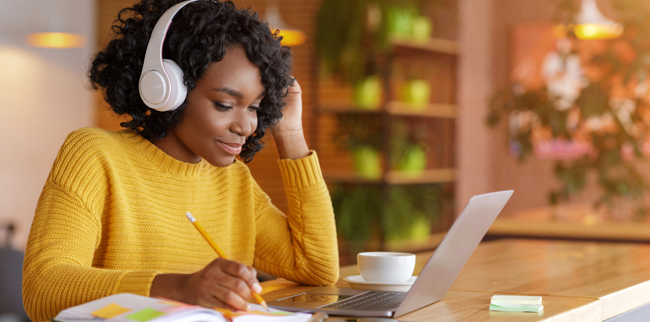 Mulher com fone de ouvido escrevendo em caderno com computador à frente