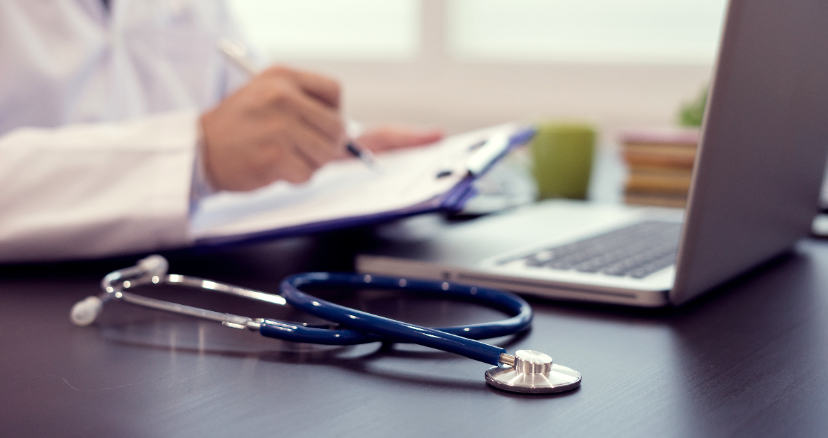 Estetoscópio em cima de mesa com médico em segundo plano segurando prancheta em frente a computador