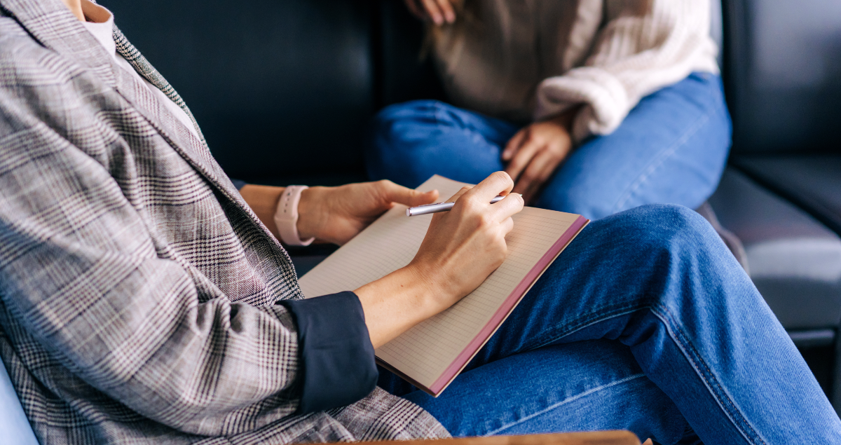 Pessoa sentada com caderno na mão em primeiro plano e outra pessoa sentada em sofá em segundo plano