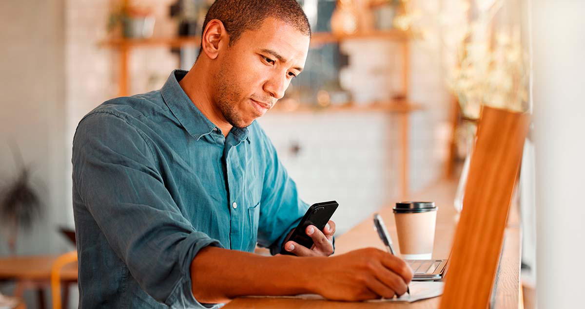 Homem escrevendo em papel com uma mão e com celular em outra mão