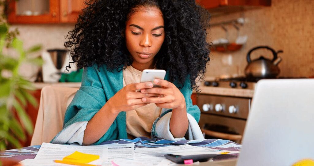 Mulher em frente a mesa com computador e papéis segurando celular nas mãos