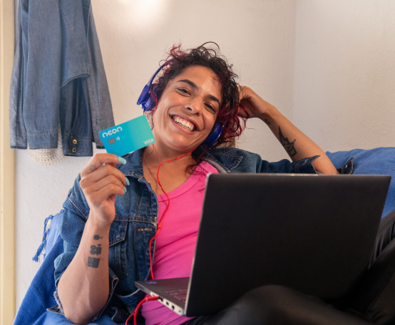 Mulher com fone de ouvido sentada em sofá com computador no colo segurando cartão Neon em uma das mãos