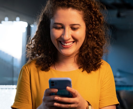 Mulher de blusa amarela sorrindo olhando para o celular em suas mãos