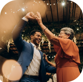 Um homem sorridente dançando com uma mulher mais velha em uma celebração. O ambiente é decorado com luzes penduradas, criando uma atmosfera festiva e alegre.