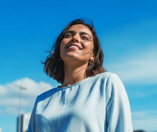 Uma jovem mulher sorrindo sob um céu azul, com cabelos soltos ao vento. Ela usa uma blusa clara e parece feliz, capturando um momento de alegria ao ar livre.