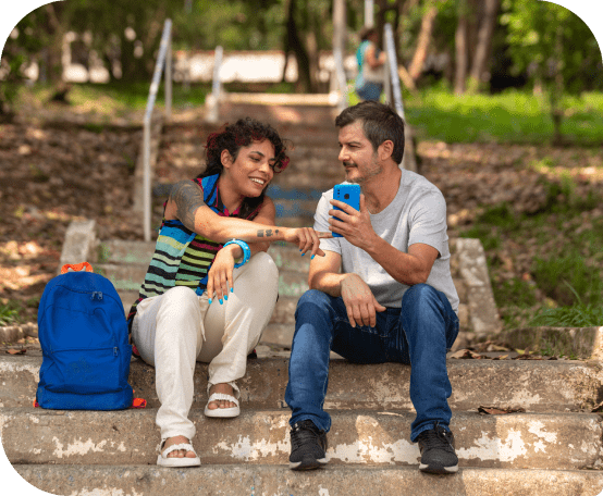 Homem e mulher sentados em escada ao ar livre, e mulher apontando para celular do homem